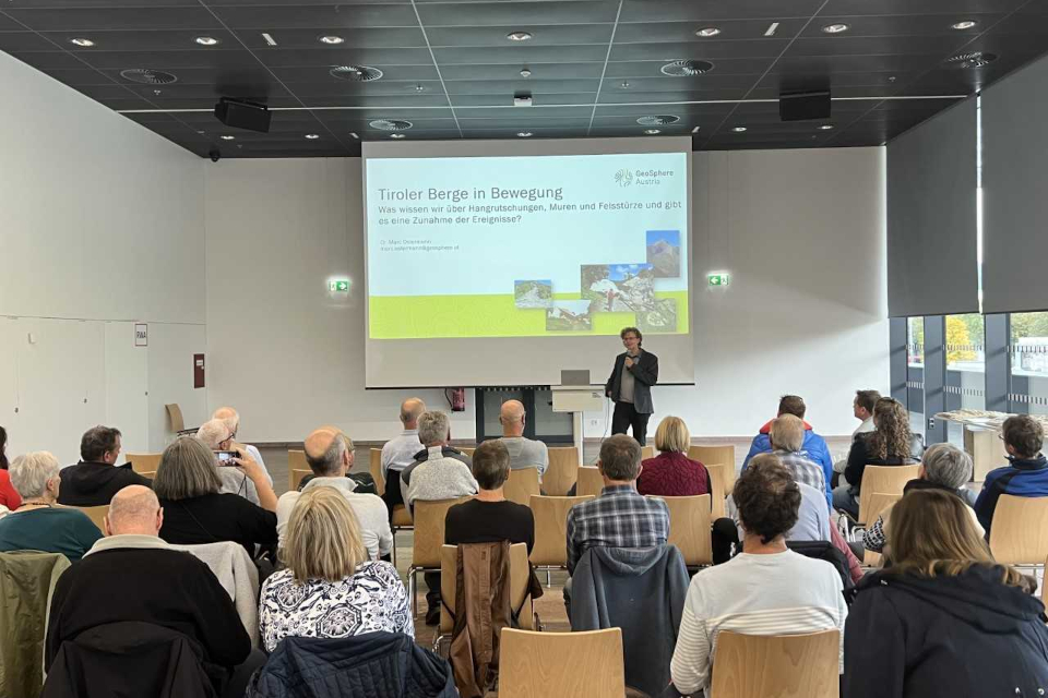 Personengruppe sitzt in einem Saal in dem ein Mann einen Vortrag hält mit einer Leinwand im Hintergrund 