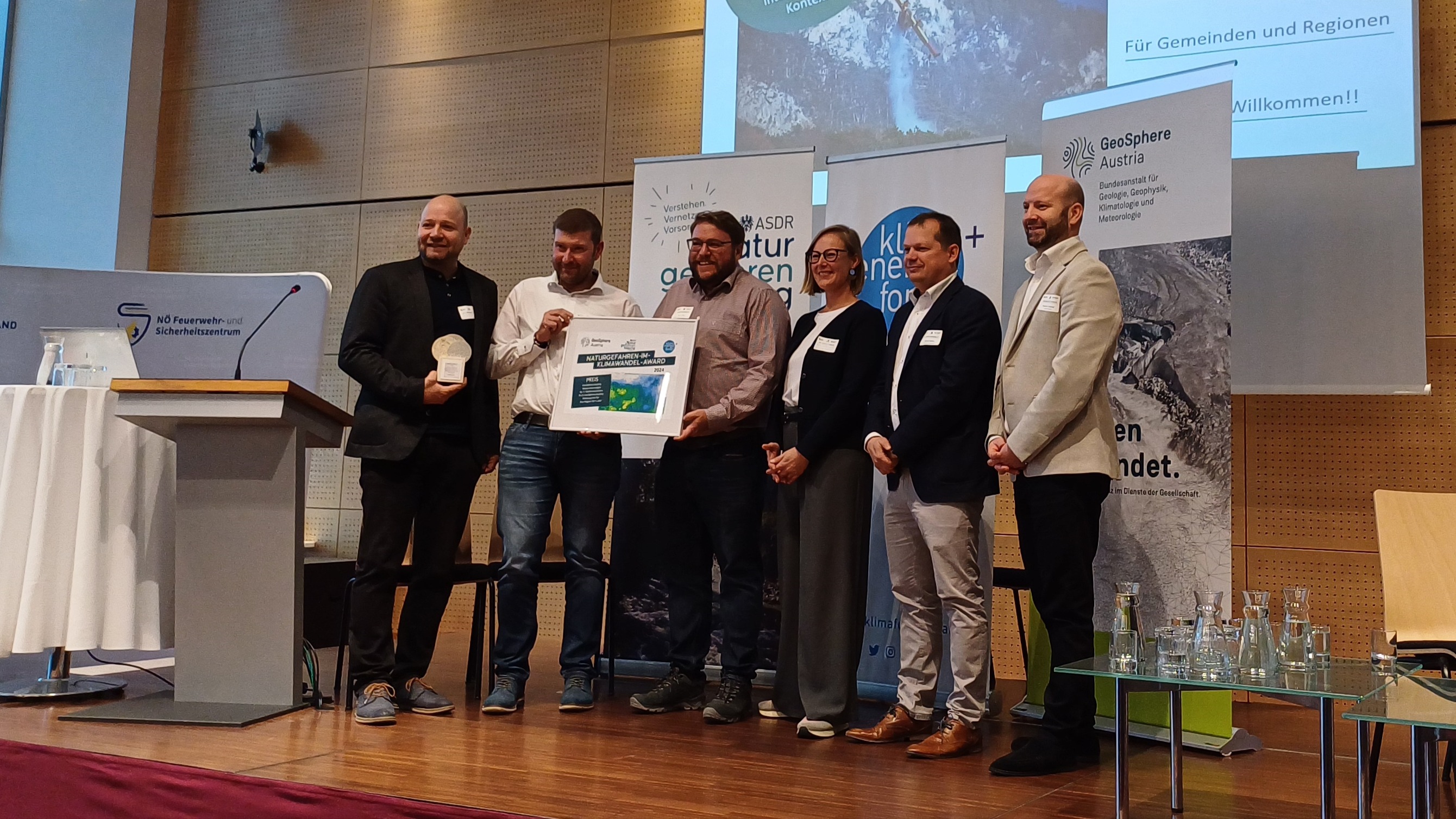 6 people stand next to each other on a podium and hold the prize in their hands. Behind them is a screen with a presentation slide.