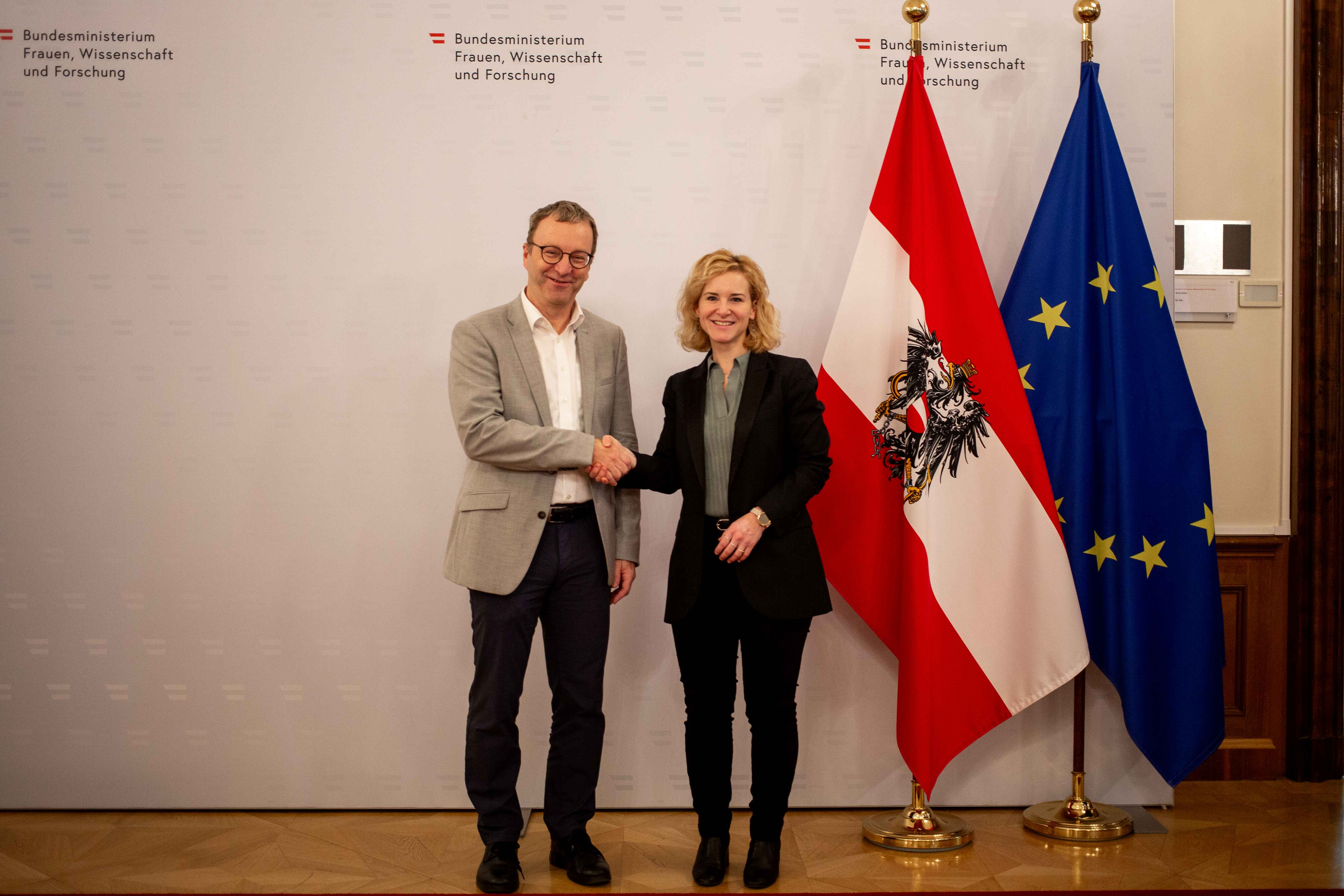 Ministerin Holzleitner gratuliert Direktor Schaffhauser neben Flaggen von Österreich und der EU