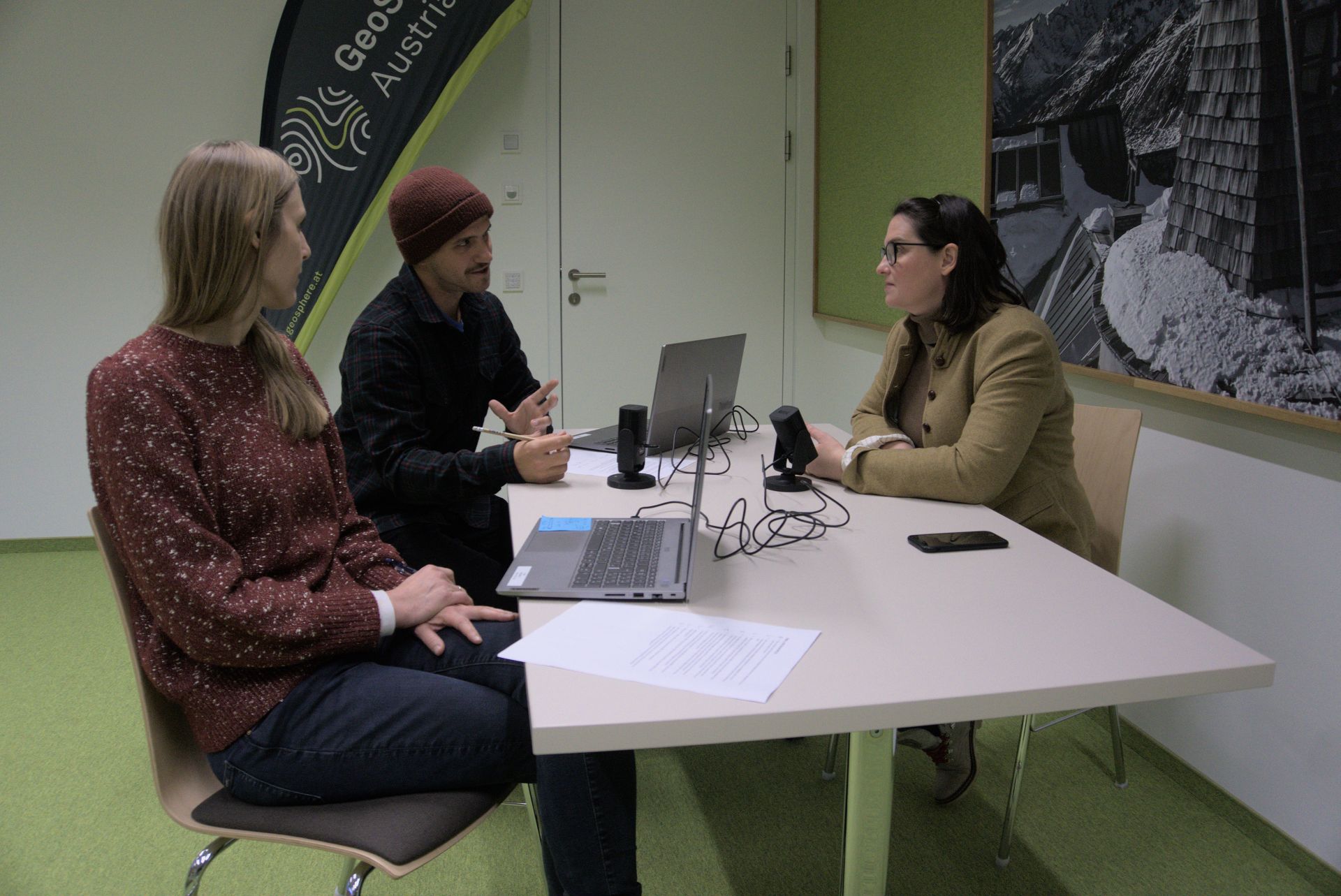 3 Personen sitzen vor einem Microphon an einem Tisch.