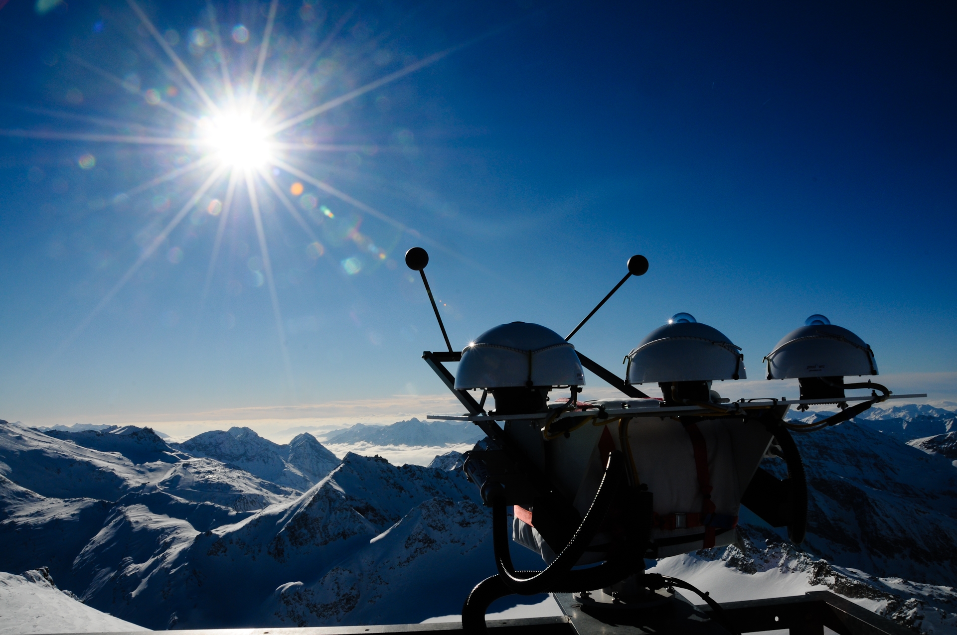 A measuring device with hemispherical superstructure and 2 antennas stands on a platform. In the background, a snow-covered mountain panorama with a blue sky and a dazzling sun with rays of sunshine.